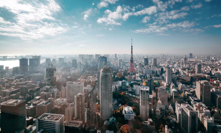 A high-angle, panoramic view of the Tokyo cityscape under a bright blue sky with scattered clouds, featuring the iconic Tokyo Tower surrounded by high-rise buildings and autumn trees.