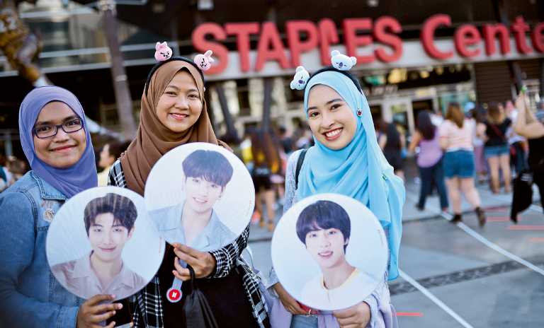 Ein Foto von drei BTS-Fans vor dem Los Angeles Staples Center. Sie halten runde Ausschnitte von drei Mitgliedern der Band in der Hand.