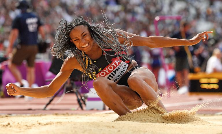 Nafi Thiam competing in the long jump with feet landing firmly in the sand and a triumphant expression on her face, hair flying about with the landing movement. Spectators and personnel blurred in the background.