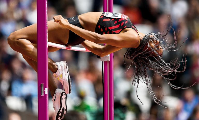 Nafi Thiam competing in the high jump, body horizontal as her back clears over the red and white pole, hair in full flow concealing her face, spectators blurred in background.