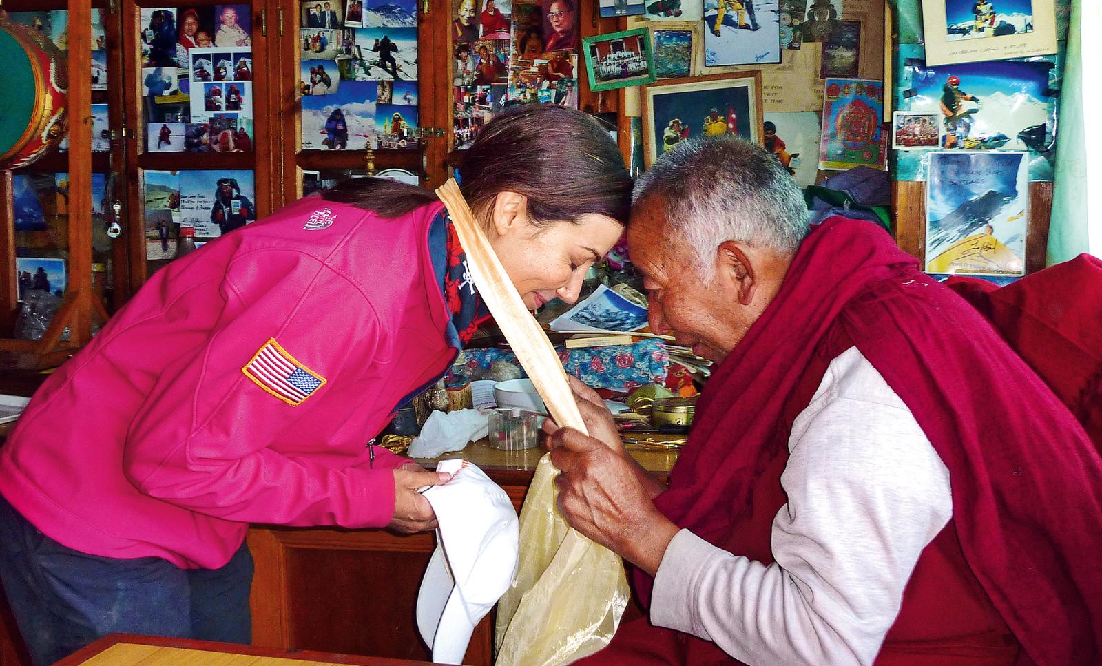 Mountain climber Alison Levine wearing a bright pink jacket bowing forward in a blessing ceremony with Lama Geshe, their heads touching, Lama wearing burgundy robe, postcards and photos on the wall.