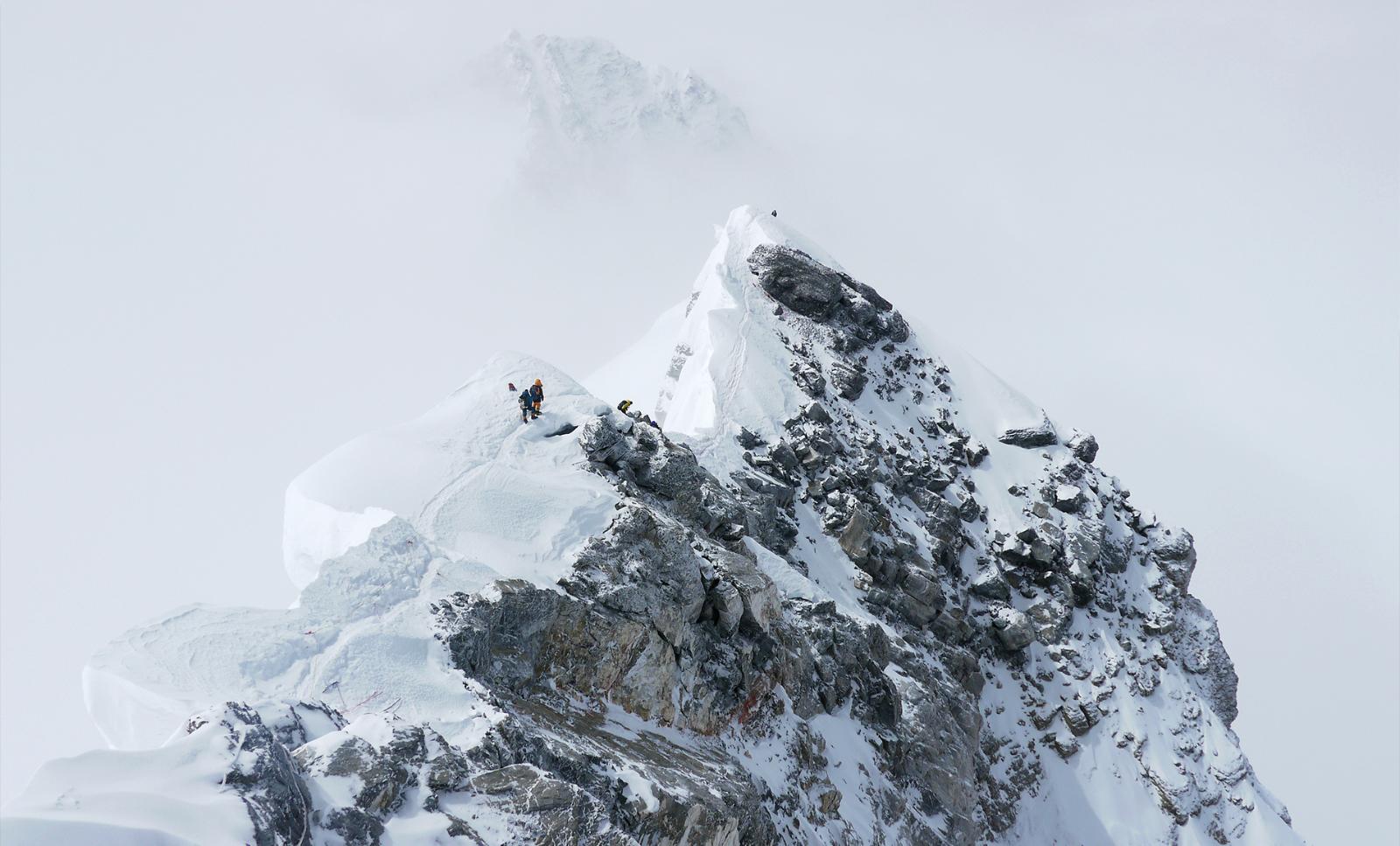 Mountain climber Laurie Bagley grinning joyfully while standing at the summit of Mount Everest, wearing a blue fleece jacket and black trousers, colorful ragged bunting and piles of stones are visible behind her.