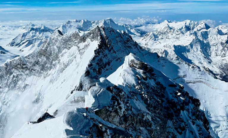 A landscape photo from above of the summit of Mount Everest on a bright sunny day, several approaching climbers visible in red outdoor clothing.