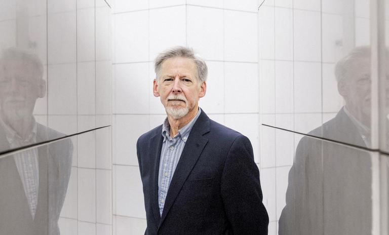 Phil Rosenzweig standing and looking into the camera, visible from the waist up, wearing a suit jacket and striped shirt, walls of shiny tiles either side of him cast his reflection.