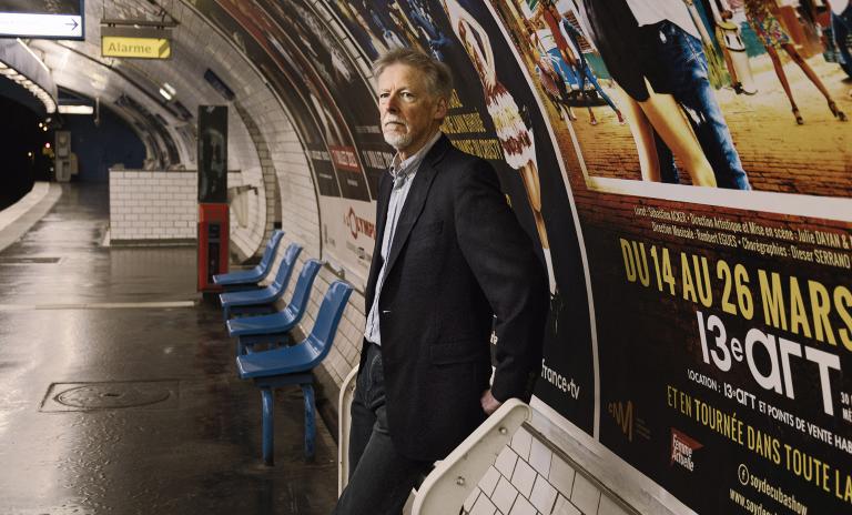 Phil Rosenzweig standing with back to the wall on a platform in the Paris underground, blue seats to his right, the tracks ahead of him, posters in French pasted on the walls behind him.