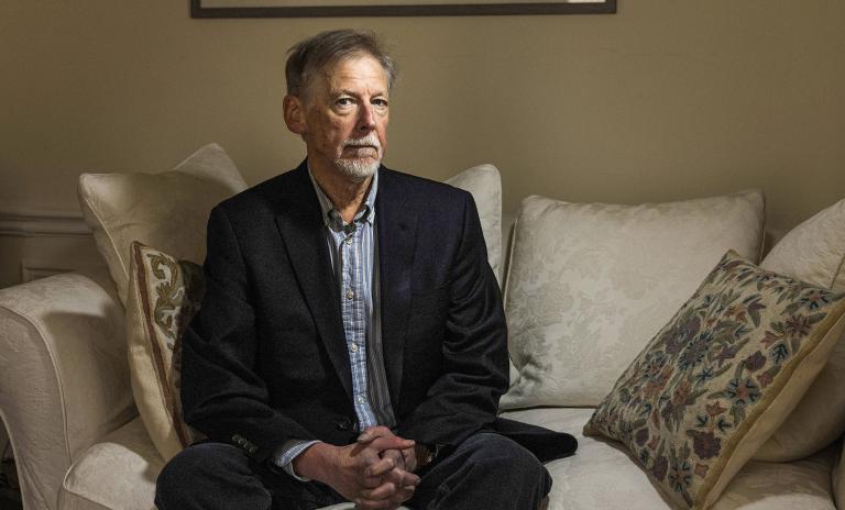 Phil Rosenzweig sitting on a cream couch, looking into the camera, serious expression, wearing a dark suit, surrounded by cushions, cream wall behind him.