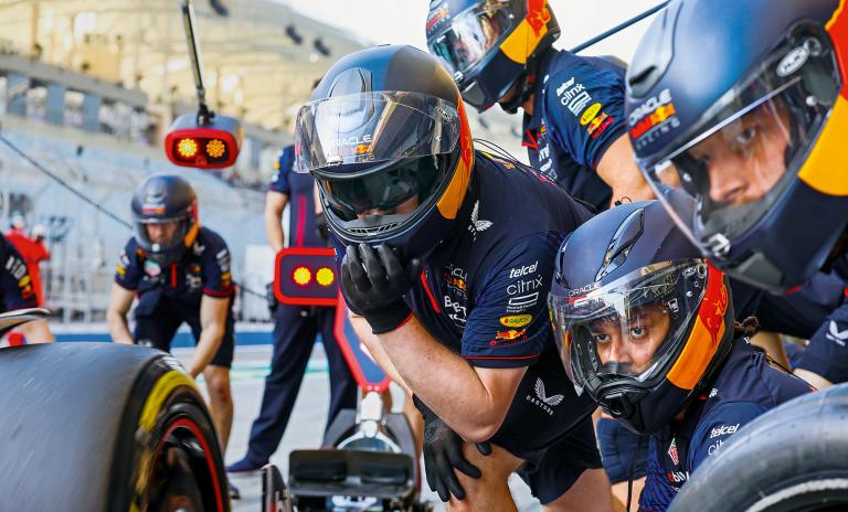 Close-up of a Formula One pit stop team, wearing helmets and sponsored clothing, poised and waiting to tend to the car at the pit stop.
