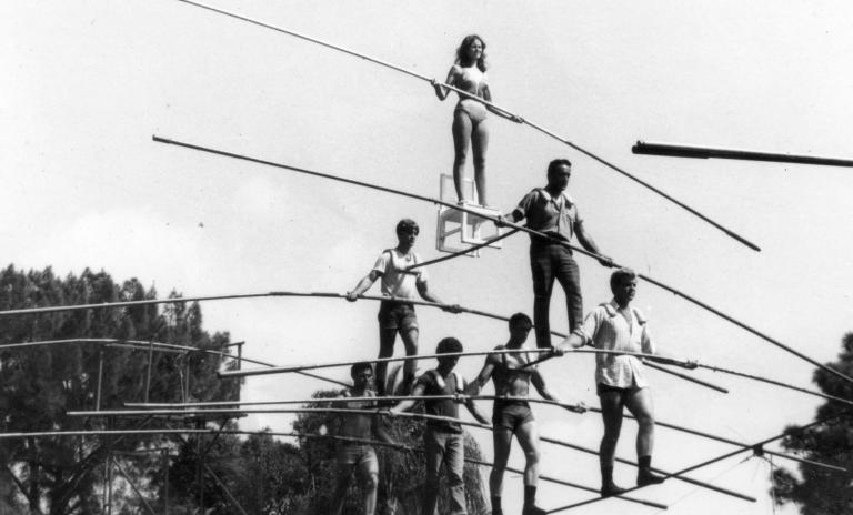 Black and white photo of high-wire acrobats performing a walk across a tightrope with long bars held in front of them for balance.