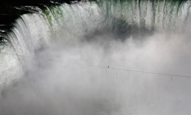 Landscape of Niagara Falls, high wire secured from left to right and a man wearing a red top and blue pants walking along the wire with arms outstretched, waterfall can be seen in the background with spray and mist rising below the wire.