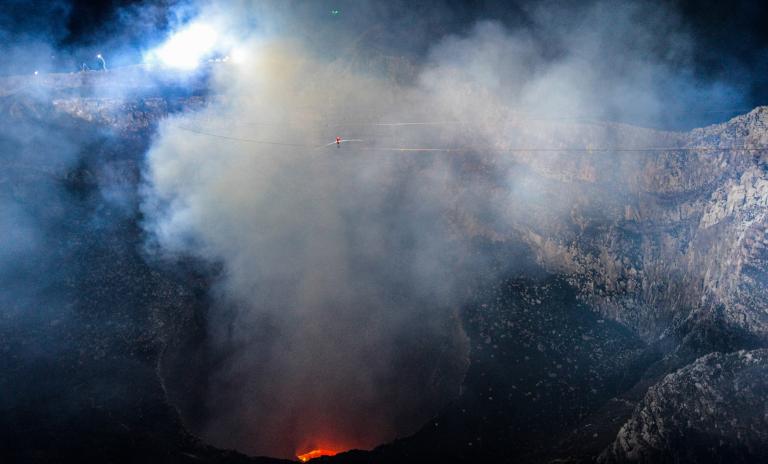 Landscape image of Masaya Volcano in Nicaragua, smoke and steam rising from orange lava at the base, high wire secured from left to right and man wearing a red top and blue pants walking along the wire through the steam and smoke, holding a long pole in front of him for balance.
