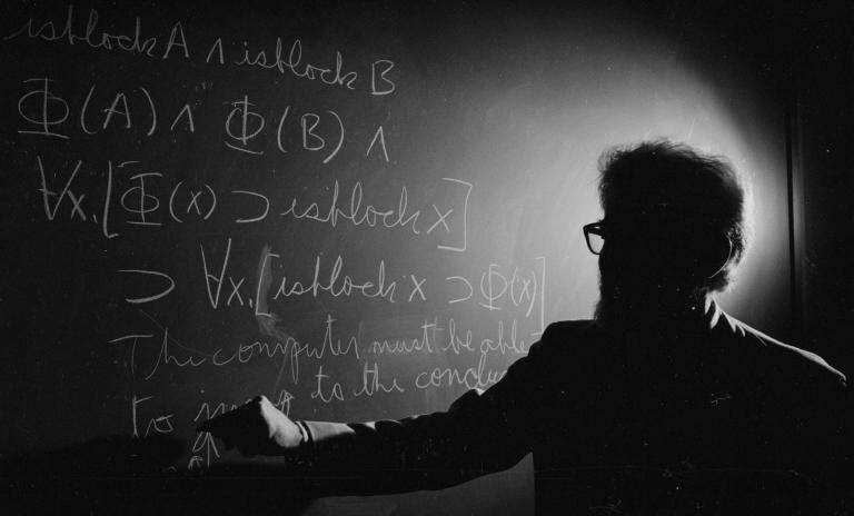 Male computer scientist with his back to the camera facing a blackboard and writing mathematical formulas on it.