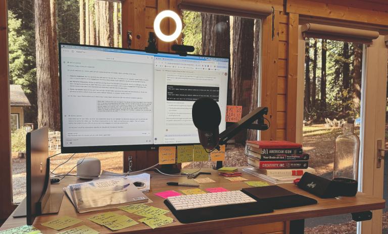 A view of Jeremy Utley’s desk in his home office showing a large computer monitor, lots of Post-it notes, a microphone, keyboard and small pile of books.