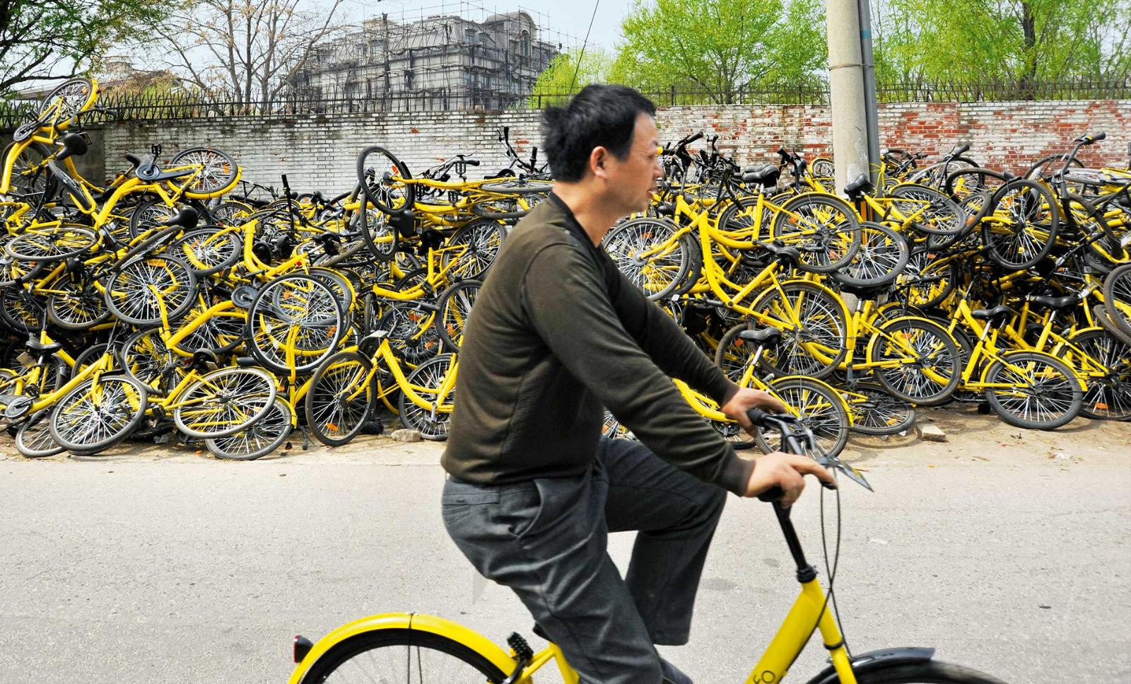 A man on an Ofo bicycle rides past a pile of discarded yellow Ofo bicyces