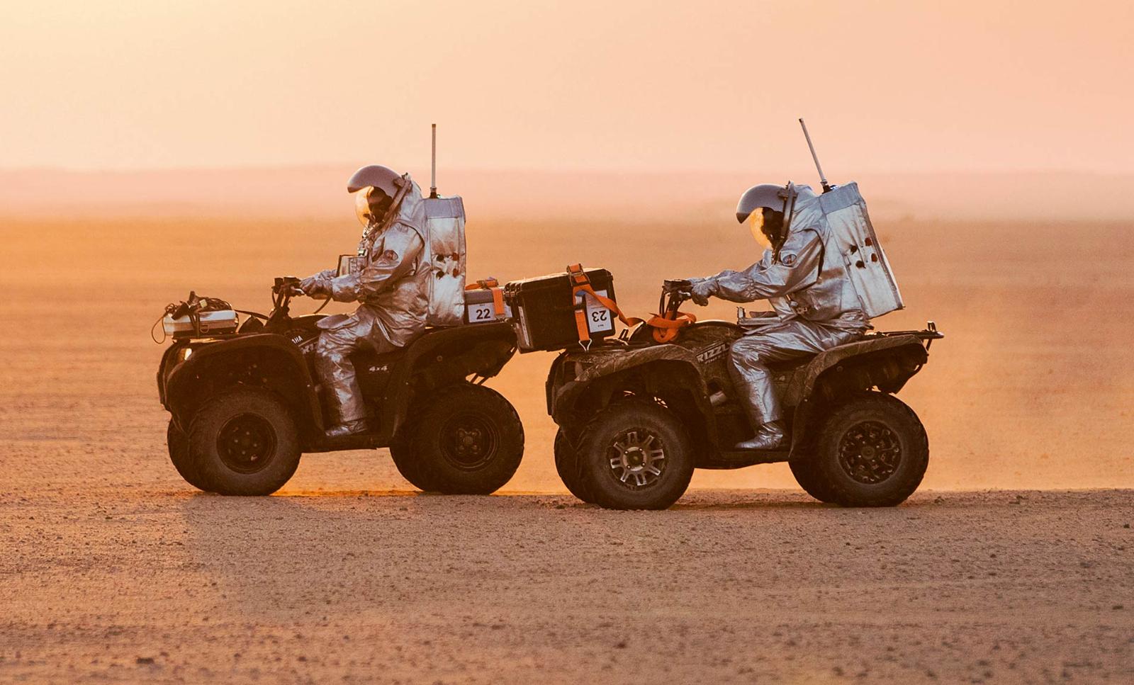 Two analog astronauts drive all-terrain vehicles across the desert in their uniform suits