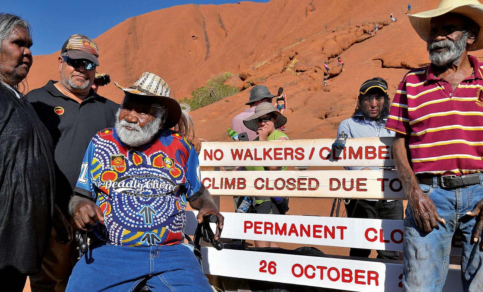 Aboriginal elders stand at the base of Uluru, also known as Ayers Rock, with a sign marking the start of a permanent climbing ban