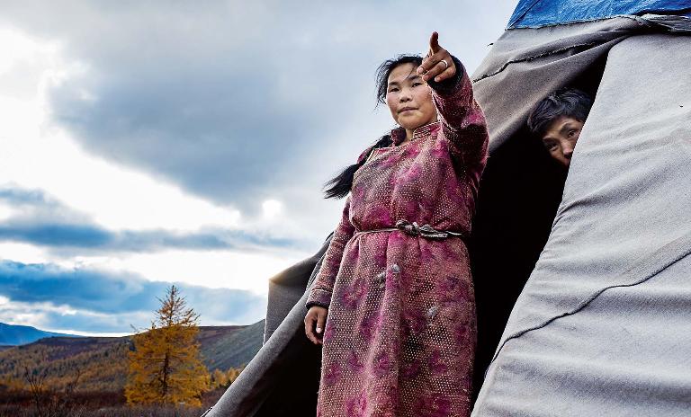 A young female member of the Mongolian Tsaatan reindeer shepherd tribe stands outside of her teepee with her husband behind her