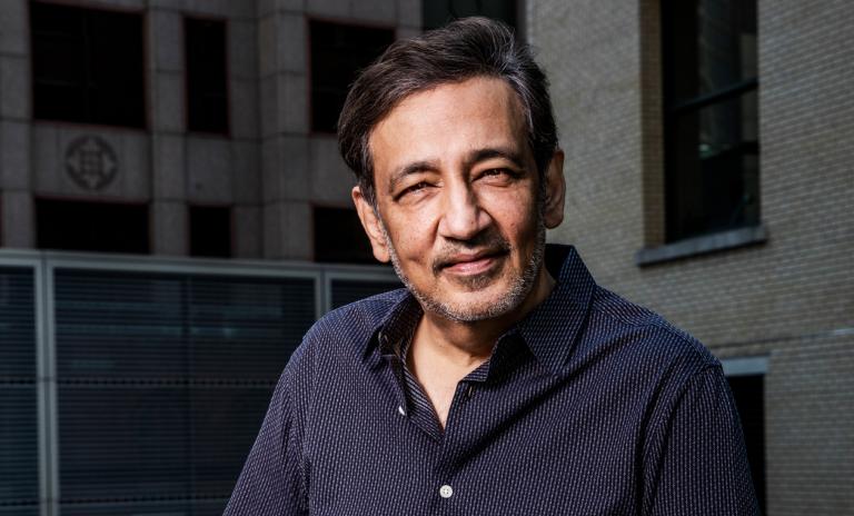 Portrait of Niren Chaudhary outdoors wearing a dark blue pinstriped shirt looking towards the camera with friendly expression. Buildings can be seen in the background behind him.