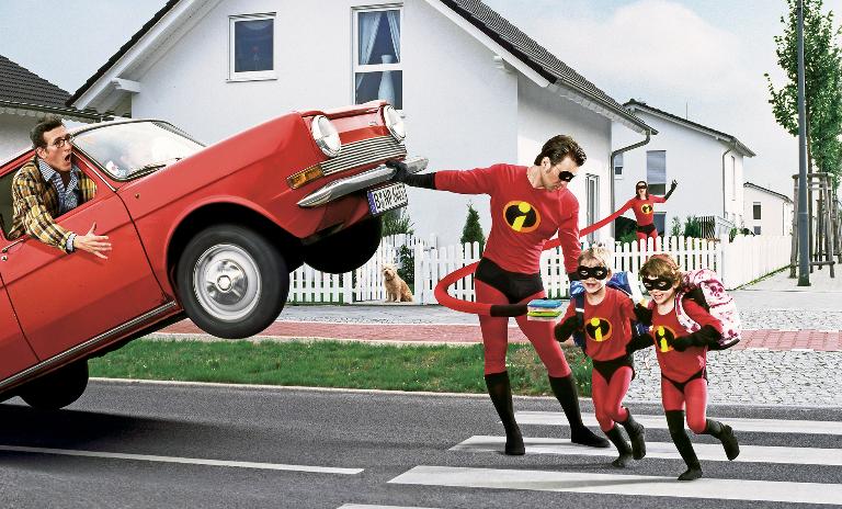 A man in a Superhero suit stands on a crosswalk and lifts a car. Schoolchildren also in Superhero suits run across the street. A woman in a Superhero suit further in the back passes them lunchboxes with her overdimensioned arm. © Maak Roberts/Gallery Stock