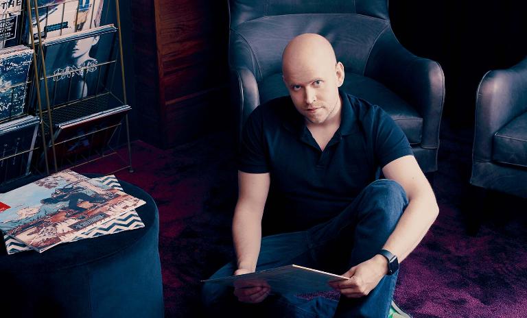 The Spotify founder and CEO, Daniel EK, sits on the floor in front of a chair, holding an album cover in his hands and looking up. (c) Wesley Mann/AUGUST