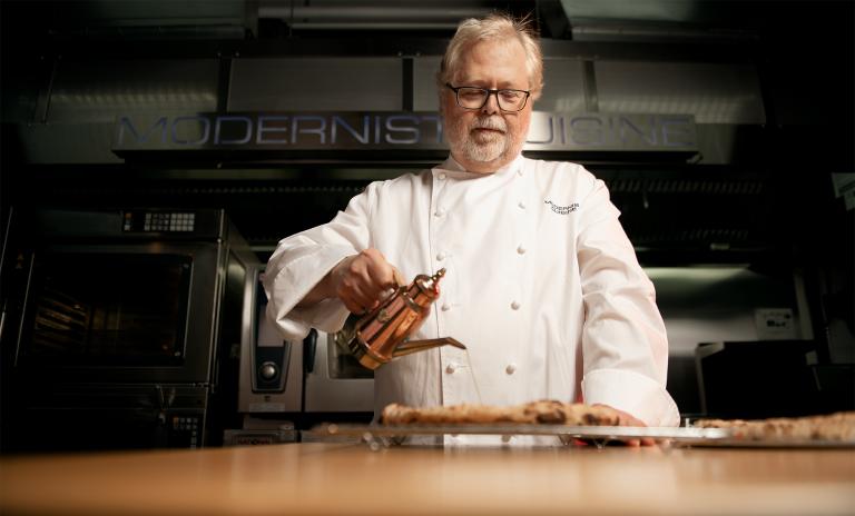 A photograph of former Microsoft CTO Nathan Myhrvold in a chef’s costume, dripping olive oil from a copper kettle onto a freshly baked pizza.