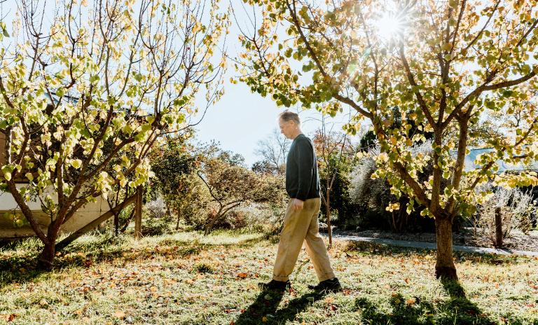 A photograph of novelist Kim Stanley Robinson walking on a lawn in the sunlight between two trees.