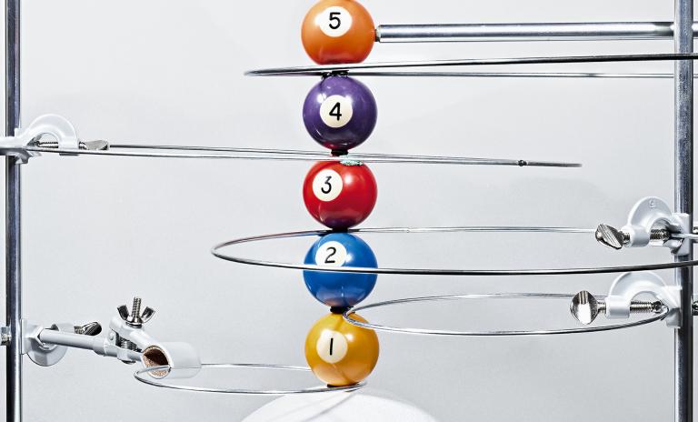 A metal contraption to represent the solar system, billiard balls numbered 1 to 5 displayed vertically from bottom to top, chrome loops encircled around each one to show its position in the solar system, held in place on a metallic frame with vice screws.