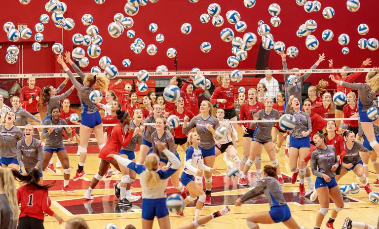 A volleyball court with multiple volleyball players in different stages of motion, appearing as if the whole court is crowded with players on both sides of the net with at least 40 volleyballs thrown into the air. A dark red interior wall of the sports hall is in the background.