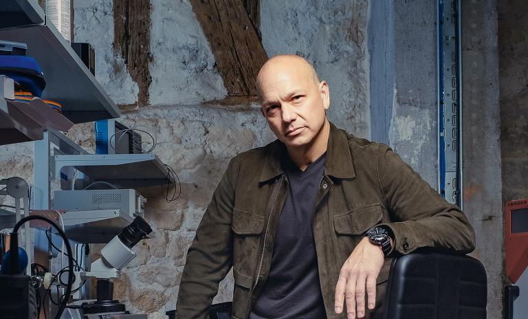 Tony Fadell looking into the camera, left arm leaning on the back of a black office chair, to his right a desk and metal shelving storing electronic equipment.