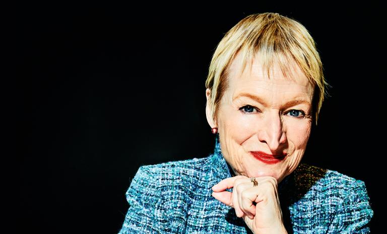 Rita Gunther McGrath sitting on a teal-colored chair wearing a woolen blazer in various shades of blue and teal, leaning on her left hand, elbow on table in front of her, facing towards camera with a friendly facial expression.