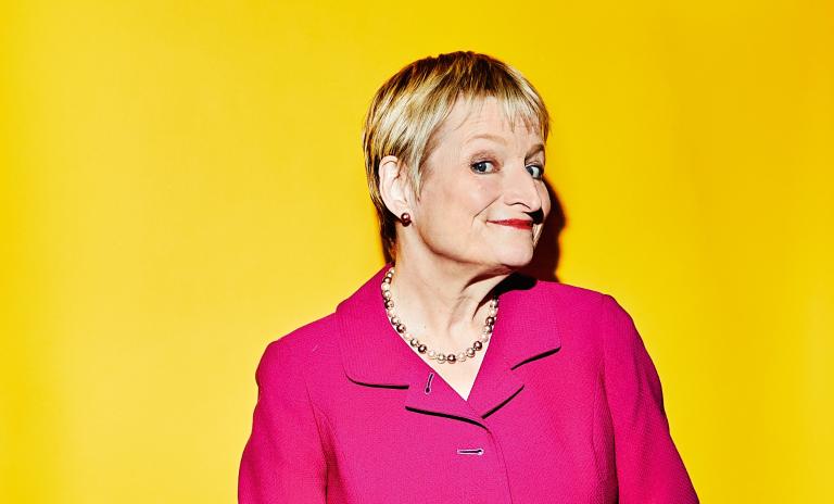 Rita Gunther McGrath wearing a bright pink blazer and black trousers, standing with her body facing towards the camera, head to her left side but eyes facing right toward the camera with a fun expression on her face.