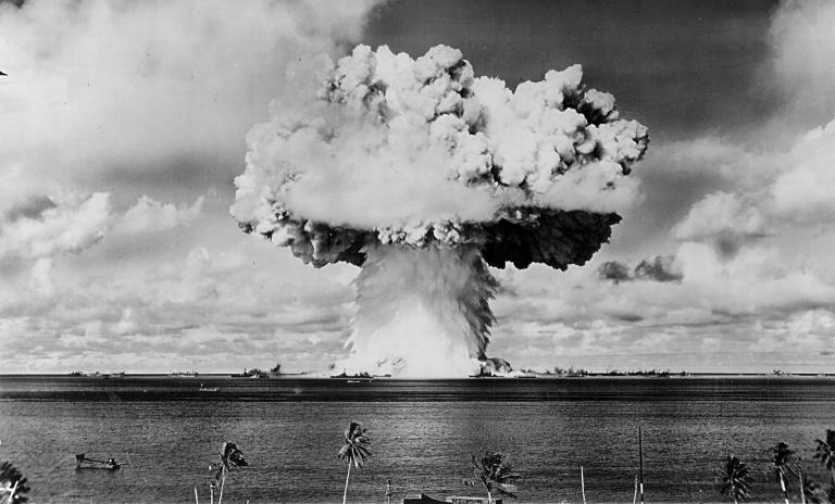 Mushroom cloud from a nuclear test explosion in the background, water, beach and palm trees in the foreground.
