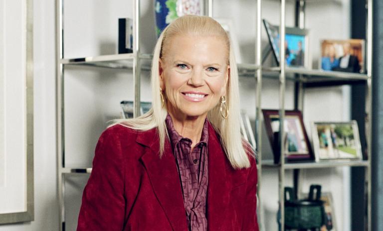 Ginni Rometty head and shoulders portrait, looking towards camera, wearing wine-colored blazer, metal office shelving behind her.