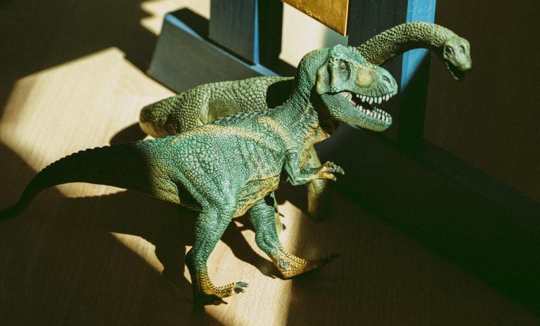 Zwei grüne Spielzeugdinosaurier – ein Tyrannosaurus und ein Diplodocus – in einem Raum mit Holzboden, beleuchtet von hereinströmendem Tageslicht.