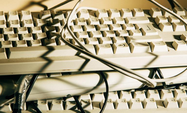 Nahaufnahme mehrerer beigefarbener Computertastaturen, um die Kabel gewickelt sind, vor einem senfgelben Hintergrund.