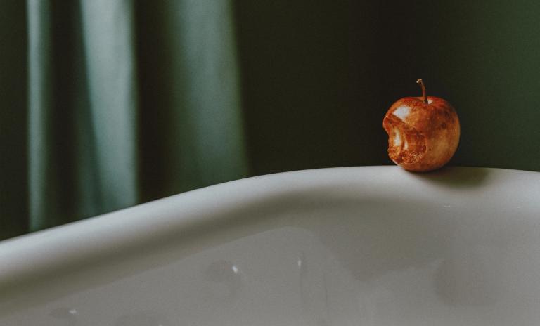 Side of a white bath with a partially eaten apple placed on its edge, a dark green shower curtain is in the background.