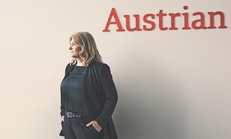Annette Mann trägt einen schwarzen Blazer und ein petrolblaues Oberteil, ihre Hände stecken in den Taschen ihrer schwarzen Hose. Sie blickt von der Kamera weg, auf der hellbeigen Wand hinter ihr ist in roten Buchstaben "Austrian" zu lesen.