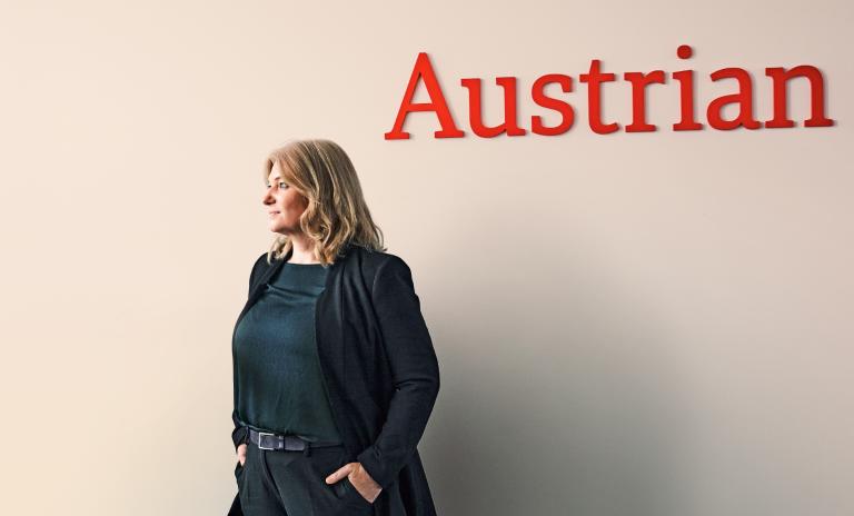 Annette Mann standing with hands in the pockets of her black trousers, also wearing a black blazer and petrol blue top, looking away from the camera, beige wall behind her and the word “Austrian” in dark red letters across the wall above her.