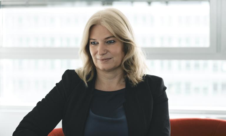 Annette Mann wearing a black blazer and trousers and a petrol blue top, sitting on a dark red sofa, looking down and away from the camera, hands clasped together in front of her, sunlight streaming through a blind at the window behind her.