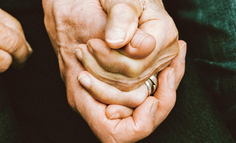 Close-up of a man’s and a woman’s hands clasped together, the woman is wearing two rings on her ring finger, dark green background.