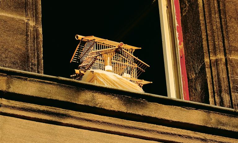 Looking up from street level at an open sash window with a small bird cage on the window ledge.