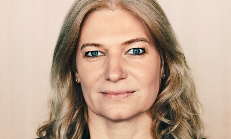 Portrait of Annette Mann wearing a black blazer and petrol blue top, looking directly into the camera with a serious but friendly expression, beige wall as a background.
