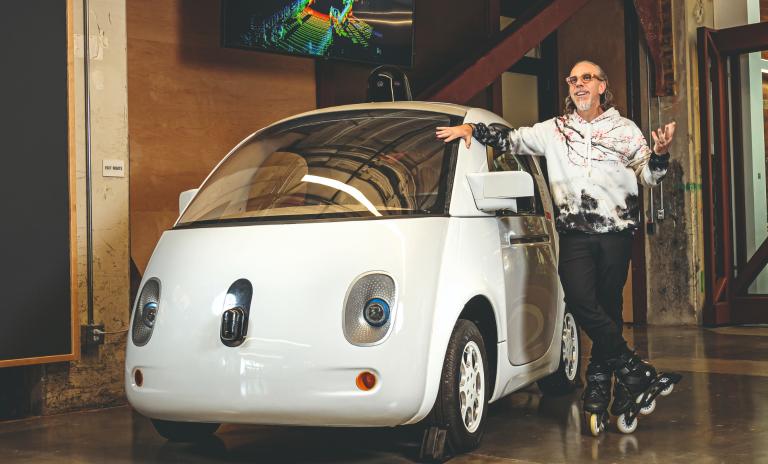 Astro Teller wearing rollerblades, a white hoodie and black pants standing next to a white Waymo car.