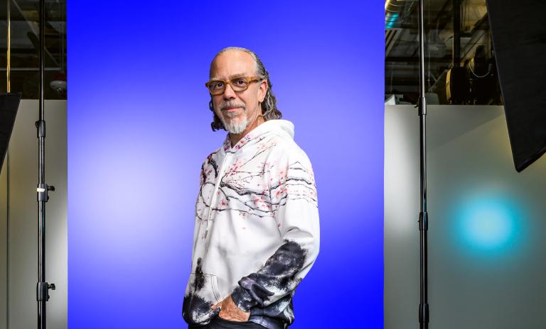 Full-length portrait of Astro Teller wearing a white hoodie and black trousers, standing sideways and twisting to face the camera.