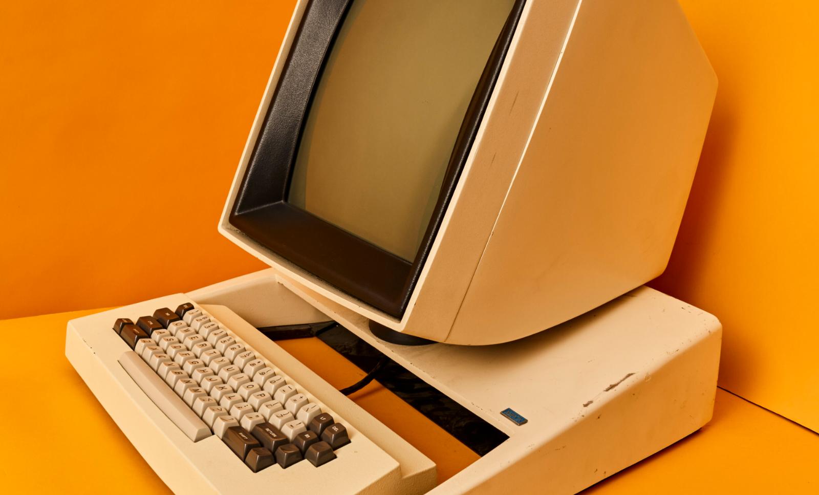 A close-up image of an old-style computer screen, keyboard and mouse attached with a cable placed in a corner with yellow-orange walls and a yellow-orange surface.