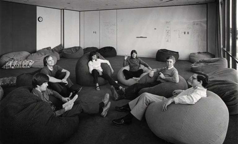 Black and white image of six people sitting on beanbags on the floor, some in office attire, others casually dressed, all sitting in relaxed positions and interacting.