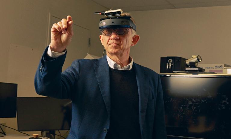Jan Vandenbrande standing and wearing virtual reality glasses, making a gesture with his right hand in relation to what he is seeing through the glasses. Screens and electrical equipment can be seen in the background.