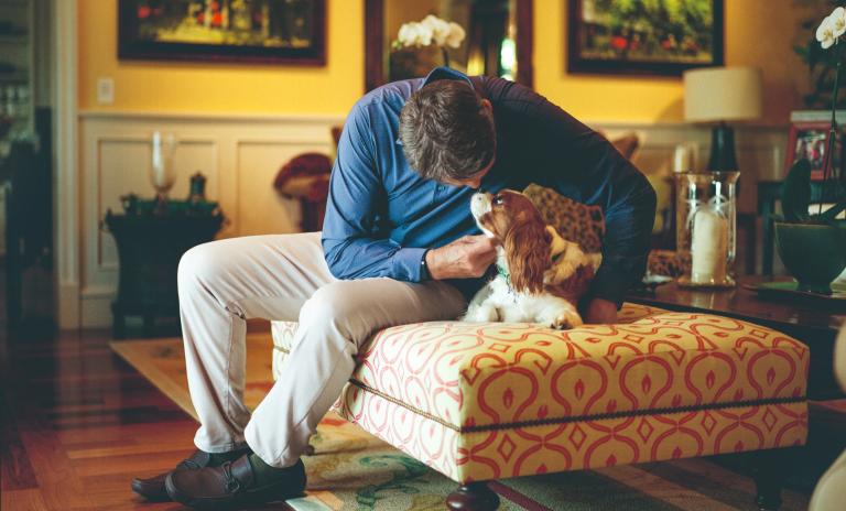 David Novak in a living room area sitting on a large foot stool and bending forwards to stroke his King Charles Spaniel who is looking up at him. The background shows golden yellow walls and hung framed paintings.
