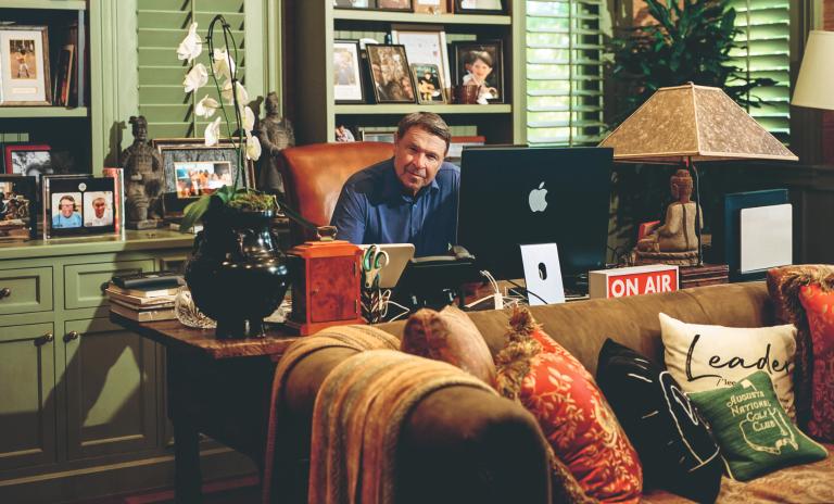 David Novak sitting on a leather chair behind his desk and looking at the camera. A green shelving unit with framed photos is behind him and a leather sofa is in front of the desk.