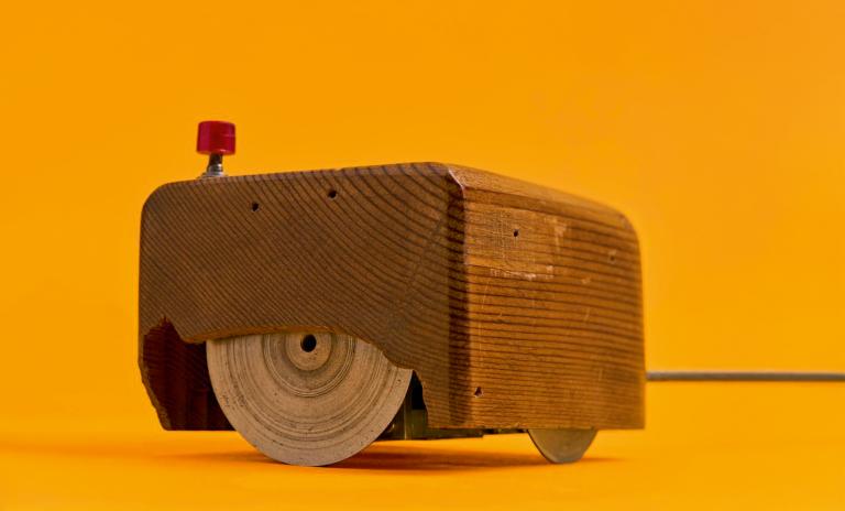 A close-up image of a wooden prototype of a computer mouse against a yellow-orange background.