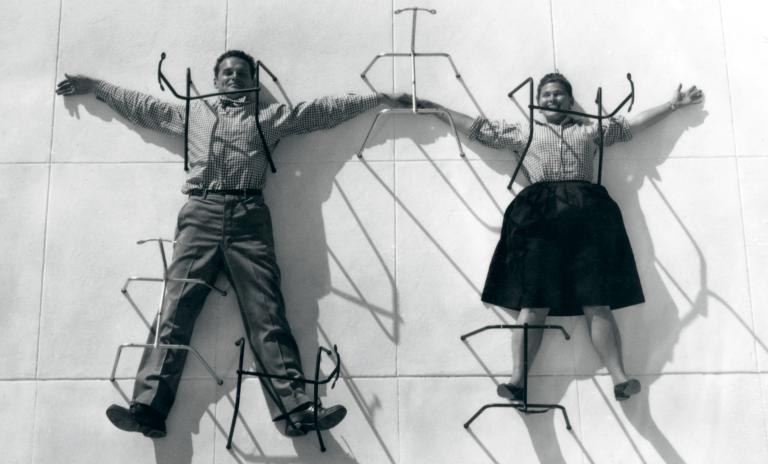 A black and white full-length photo of Charles and Ray Eames smiling with arms and legs spread out and pretending to be pinned to the ground by their own chair bases.